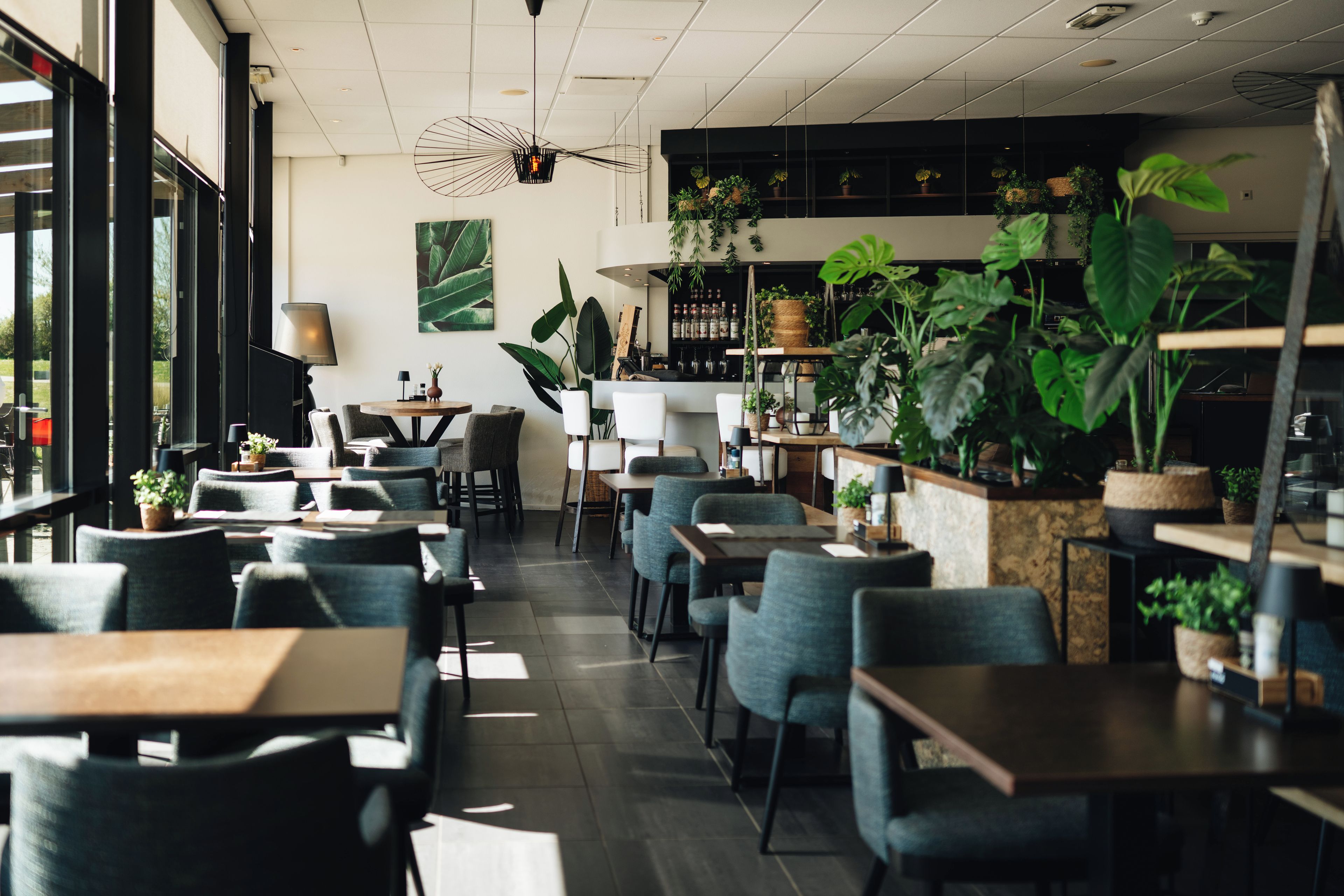 A modern restaurant interior with multiple seating areas, including tables and chairs, and a bar area with shelves of bottles and potted plants.