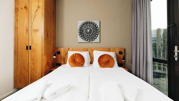 A double bed with white linens, two orange accent pillows, a wooden headboard, a wall-mounted circular artwork, and a window with curtains.