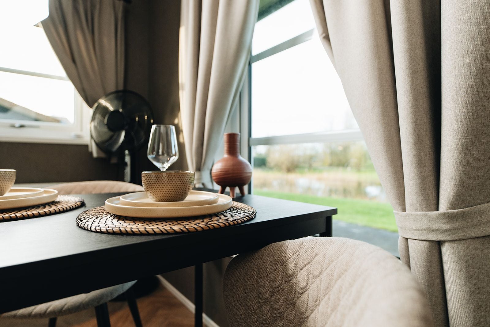 Una mesa de comedor con una copa de vino, un bol y un florero, frente a una ventana con cortinas.