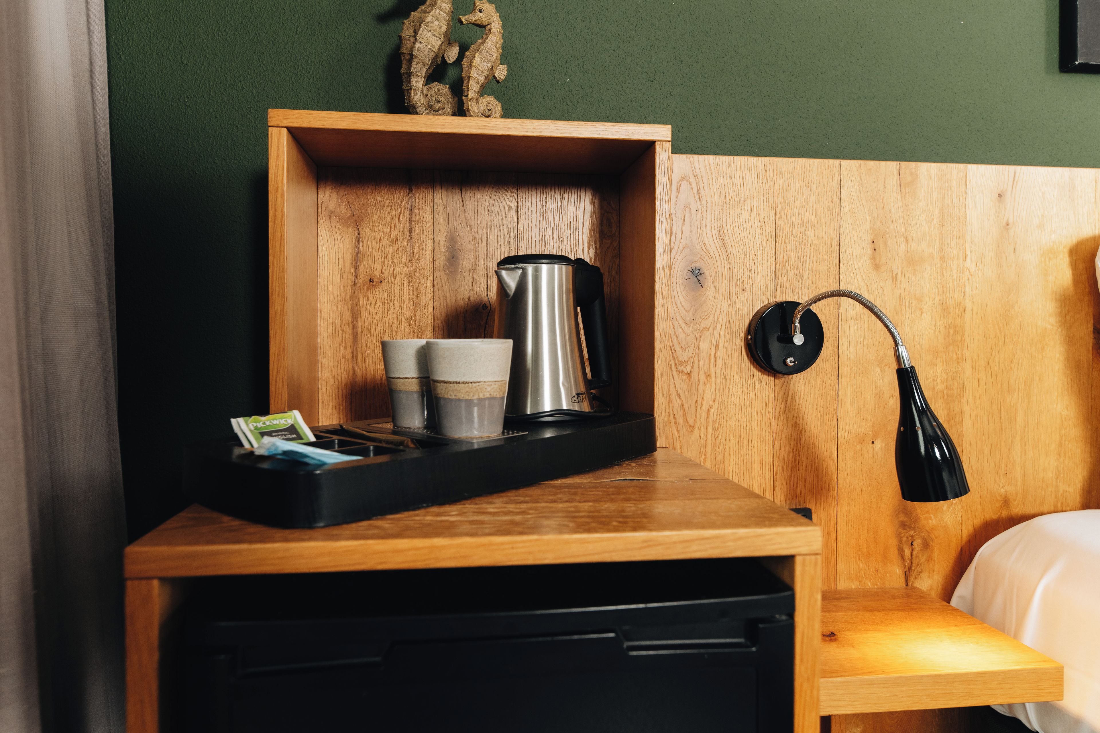 A wooden bedside table with a black tray holding a coffee pot, cups, and a pack of tissues.