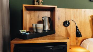 A wooden bedside table with a black tray holding a coffee pot, cups, and a pack of tissues.