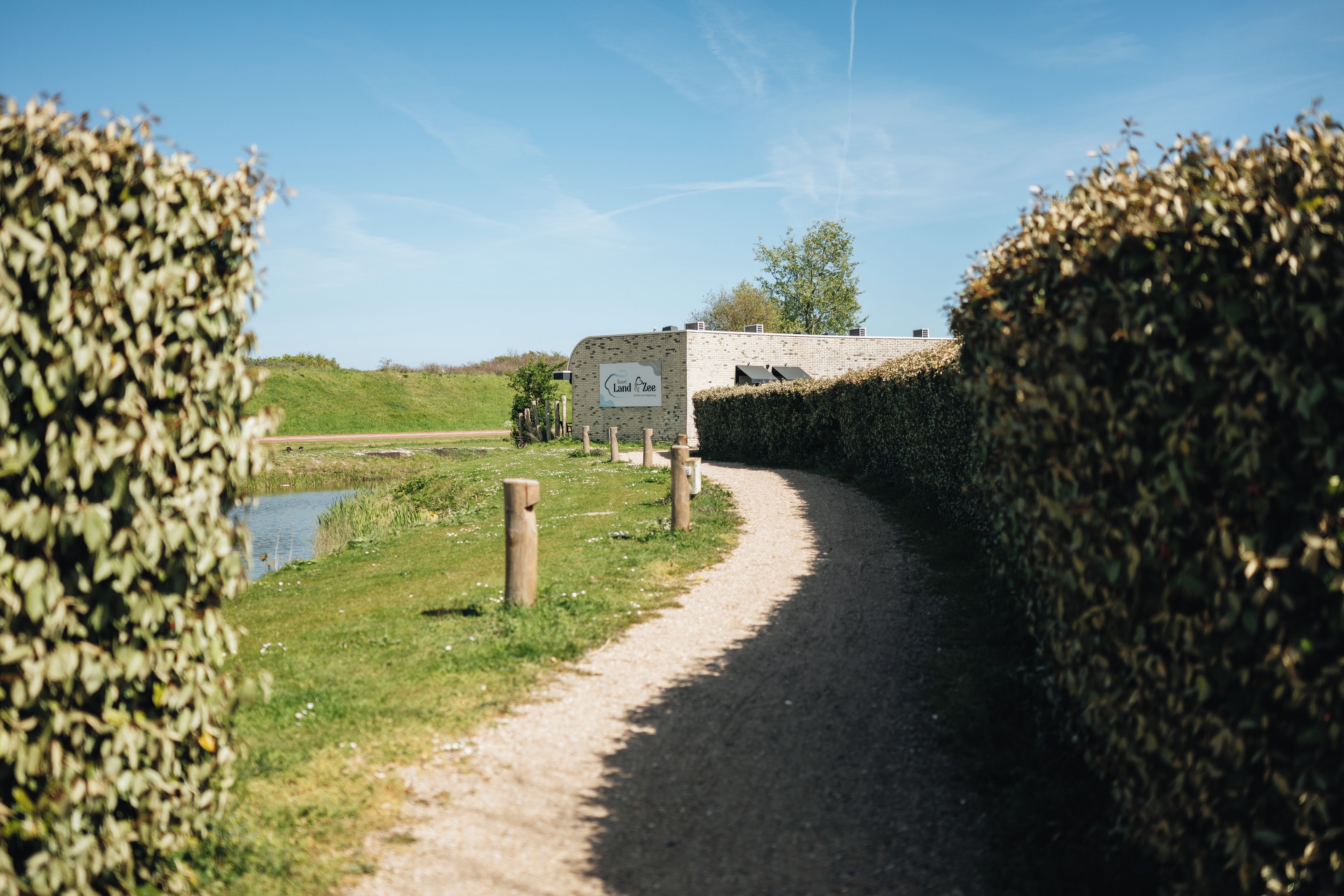 Un chemin de gravier mène à un bâtiment sur lequel est apposée une enseigne. L’ensemble est entouré de verdure et bordé par un plan d’eau.