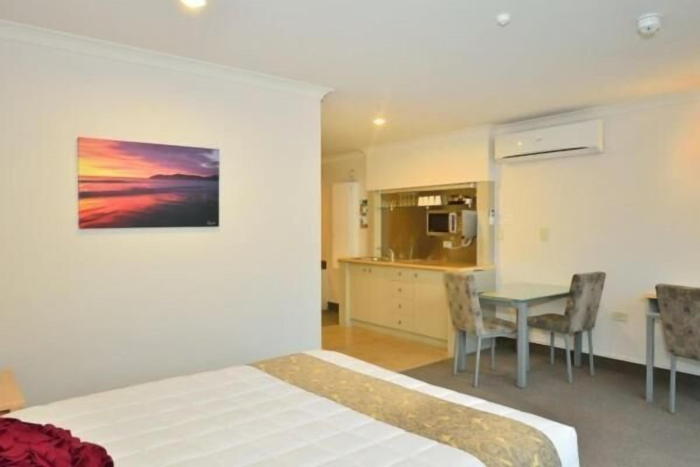 Quarto de hotel com cama, pintura de pôr do sol, cozinha compacta e área de refeições com cadeiras.