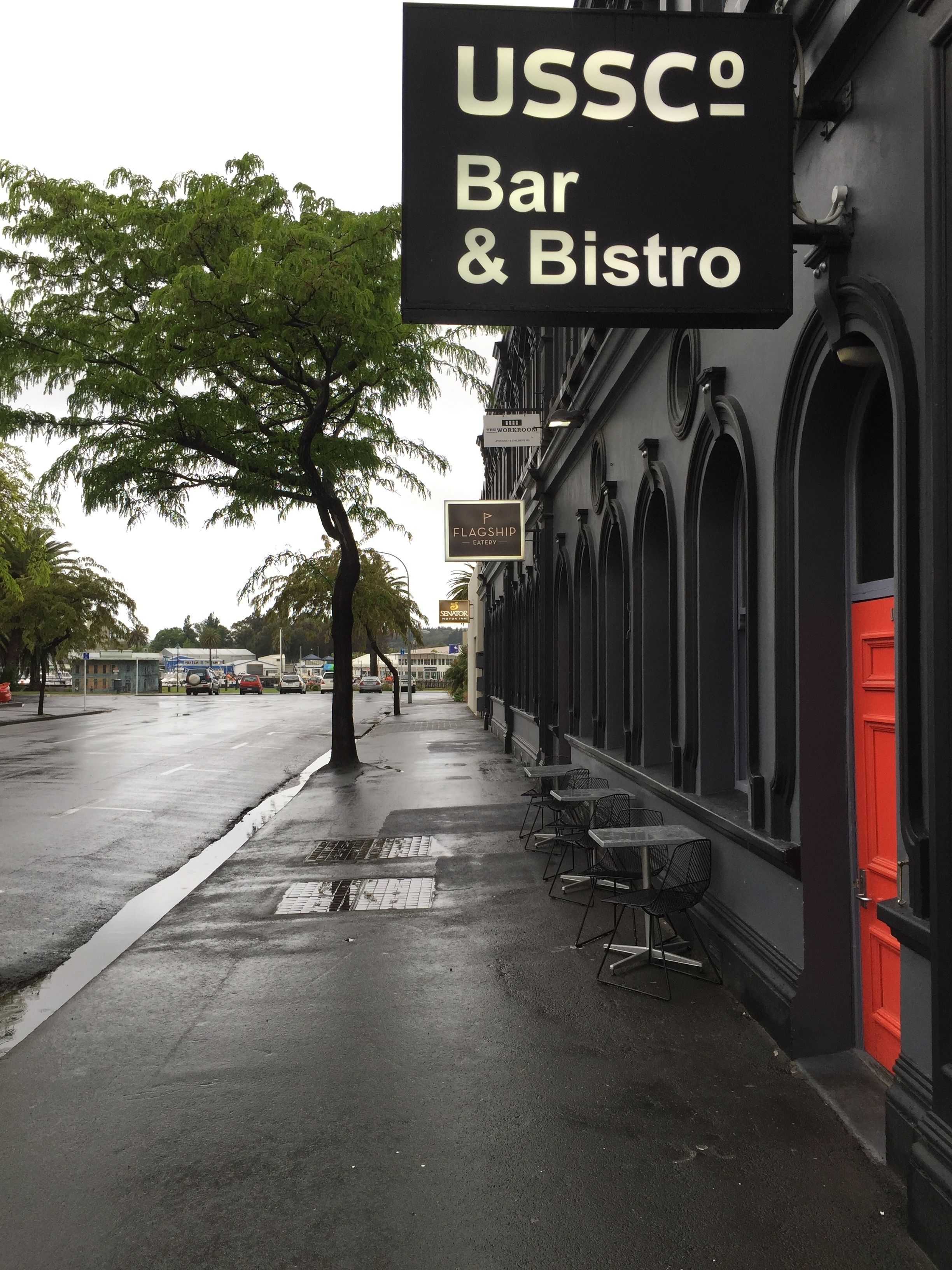 A sidewalk with outdoor seating, a sign for USSCO Bar & Bistro, and a building with arched windows.