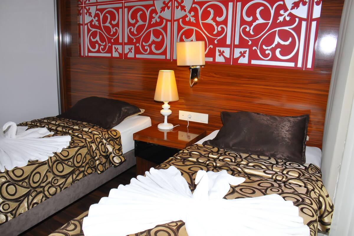 A hotel room with two beds, each with a patterned bedspread and pillows, a bedside table with a lamp, and a decorative wall panel.