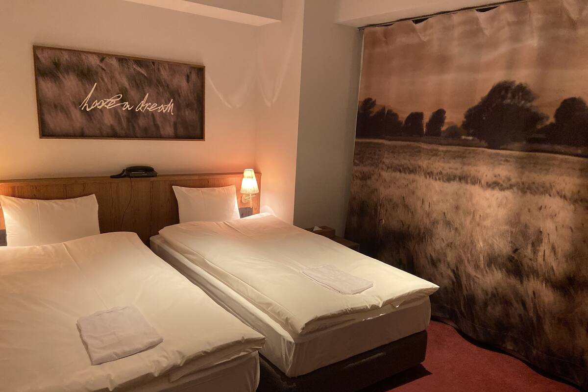 A hotel room with two beds, a headboard, a bedside lamp, a wall-mounted artwork, and a large window with a view of a field.
