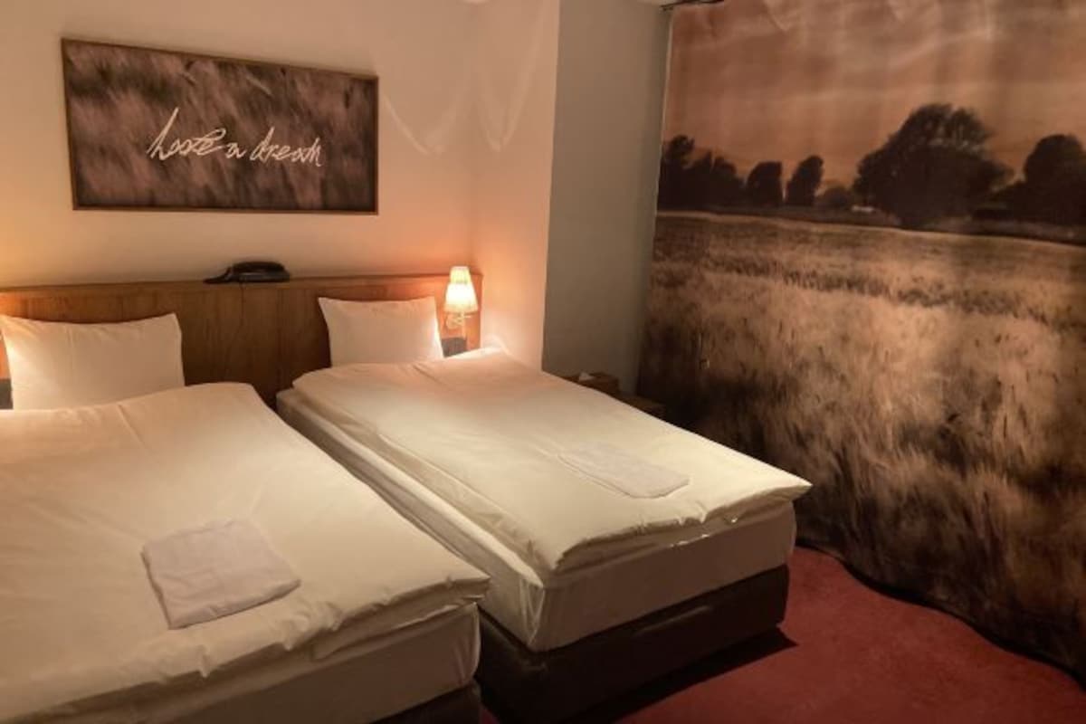 A hotel room with two beds, a headboard, a wall-mounted artwork with the phrase "Have a dream," and a large tapestry depicting a landscape.