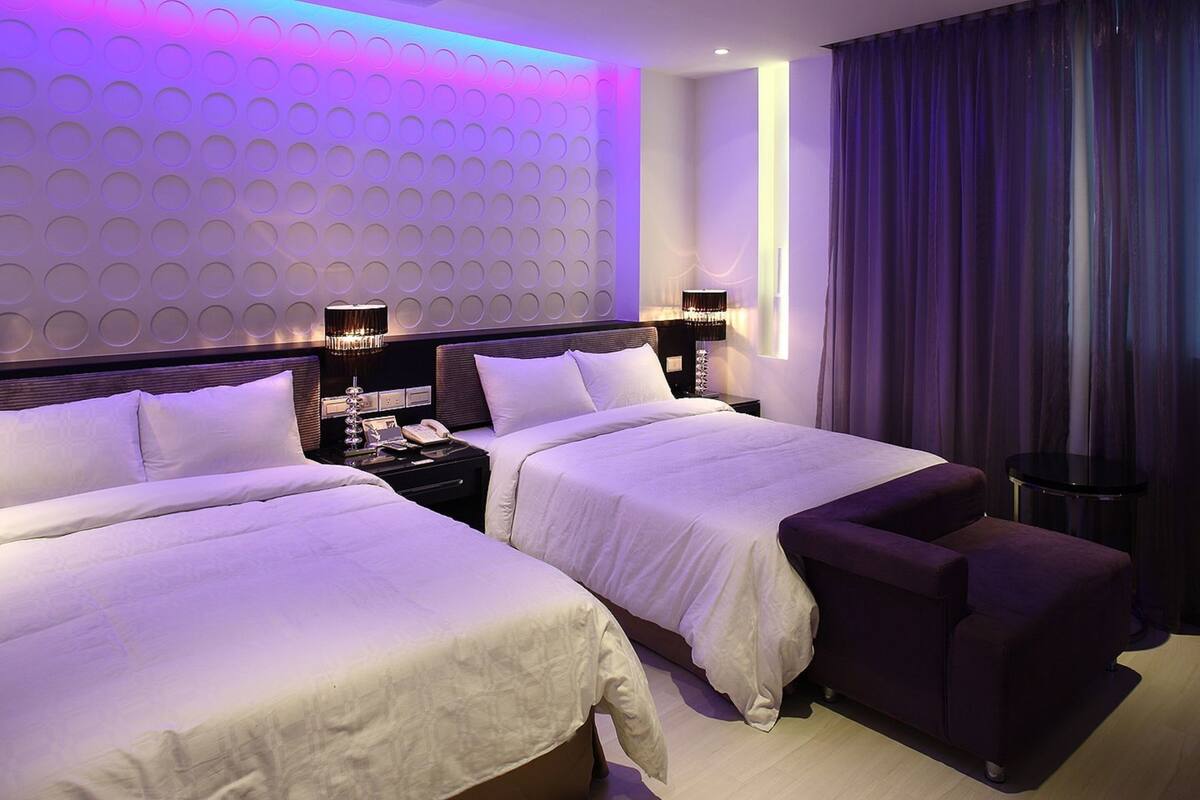 A hotel room with two beds, a nightstand, a lamp, and a view of the outside.