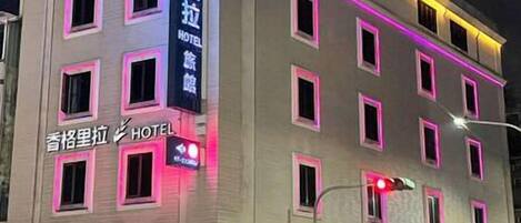 Un hotel de varios pisos con letreros iluminados, entrada visible y una zona de estacionamiento con vehículos.