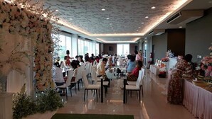 A spacious indoor dining area with round tables and chairs, large windows, and a decorative floral arrangement on the left. - The Sila Sriracha (Si Racha)