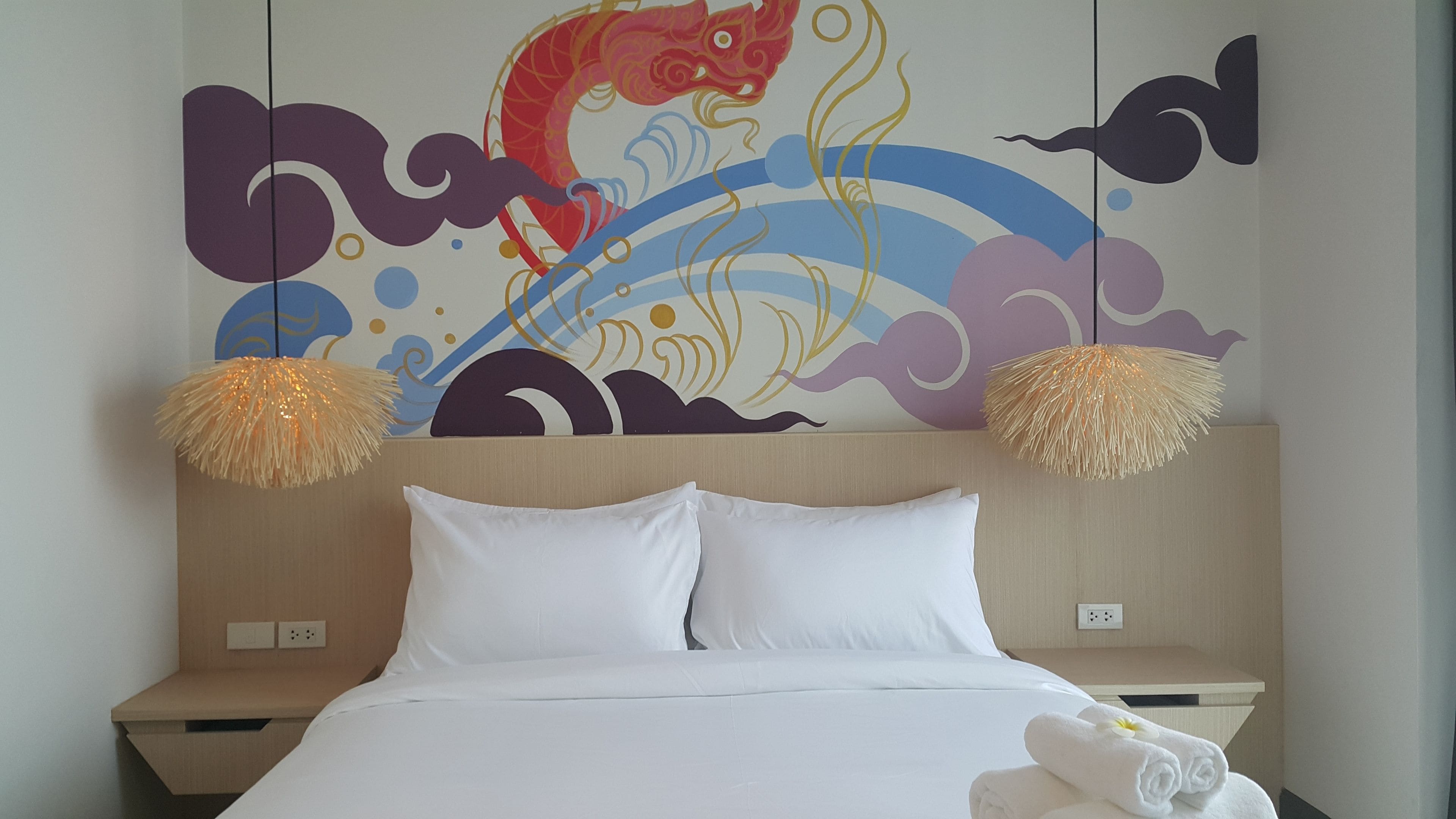 Une chambre d’hôtel avec un lit, des oreillers blancs et une fresque murale décorative. Il y a deux tables de chevet avec des lampes.