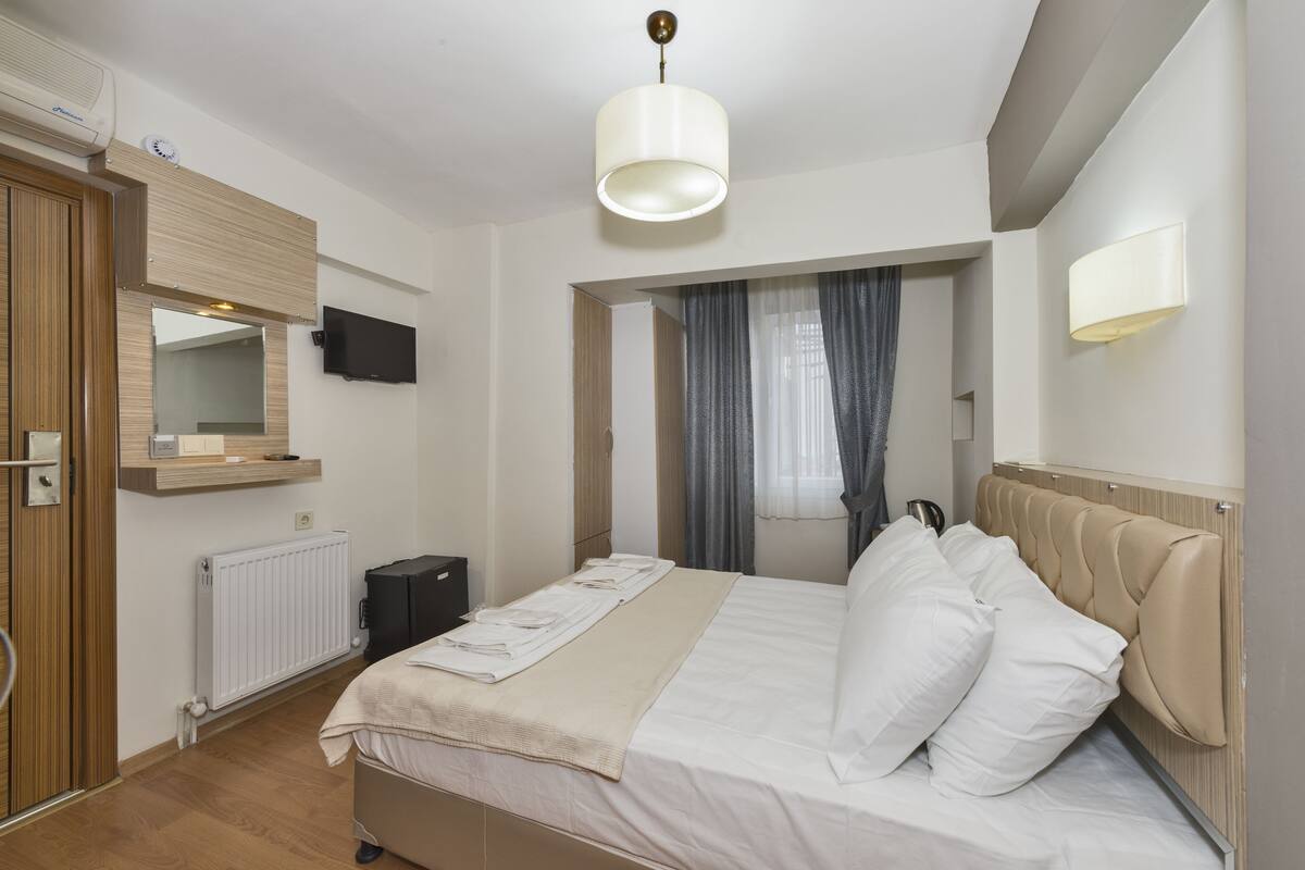Una habitación de hotel con cama, televisor, espejo, radiador y ventana con cortinas.
