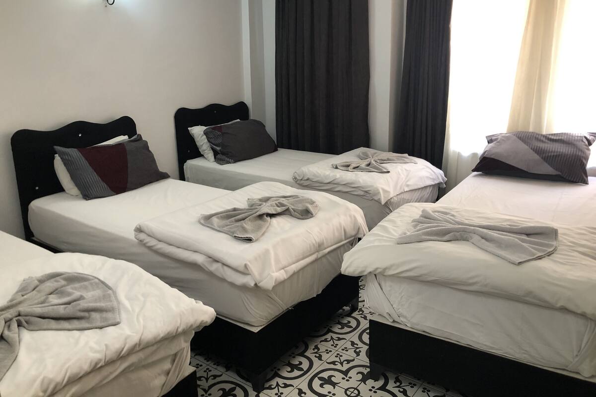 A hotel room with four single beds, each with a white bedspread and a towel. There is a black headboard on the left, and a wall-mounted light fixture above the bed on the right.