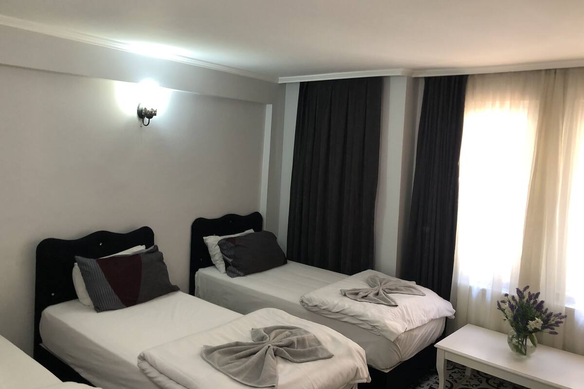 A hotel room with two single beds, a small bedside table with a vase of flowers, and a window with curtains.