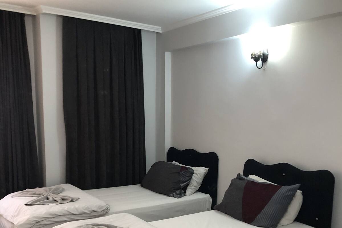 A hotel room with three beds, each with a towel on the bed. The room has a patterned floor and a light fixture on the wall.