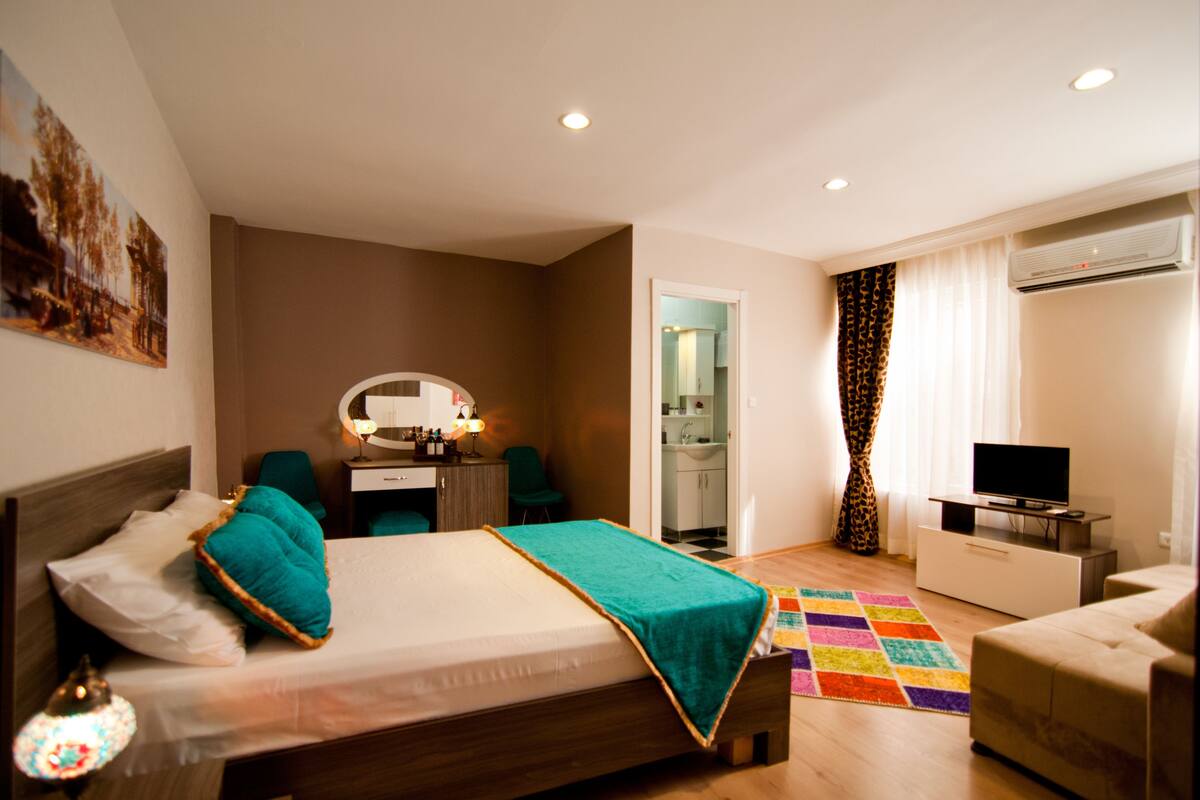 A hotel room with a bed, a desk, a chair, a TV, an air conditioning unit, and a bathroom visible through an open door.