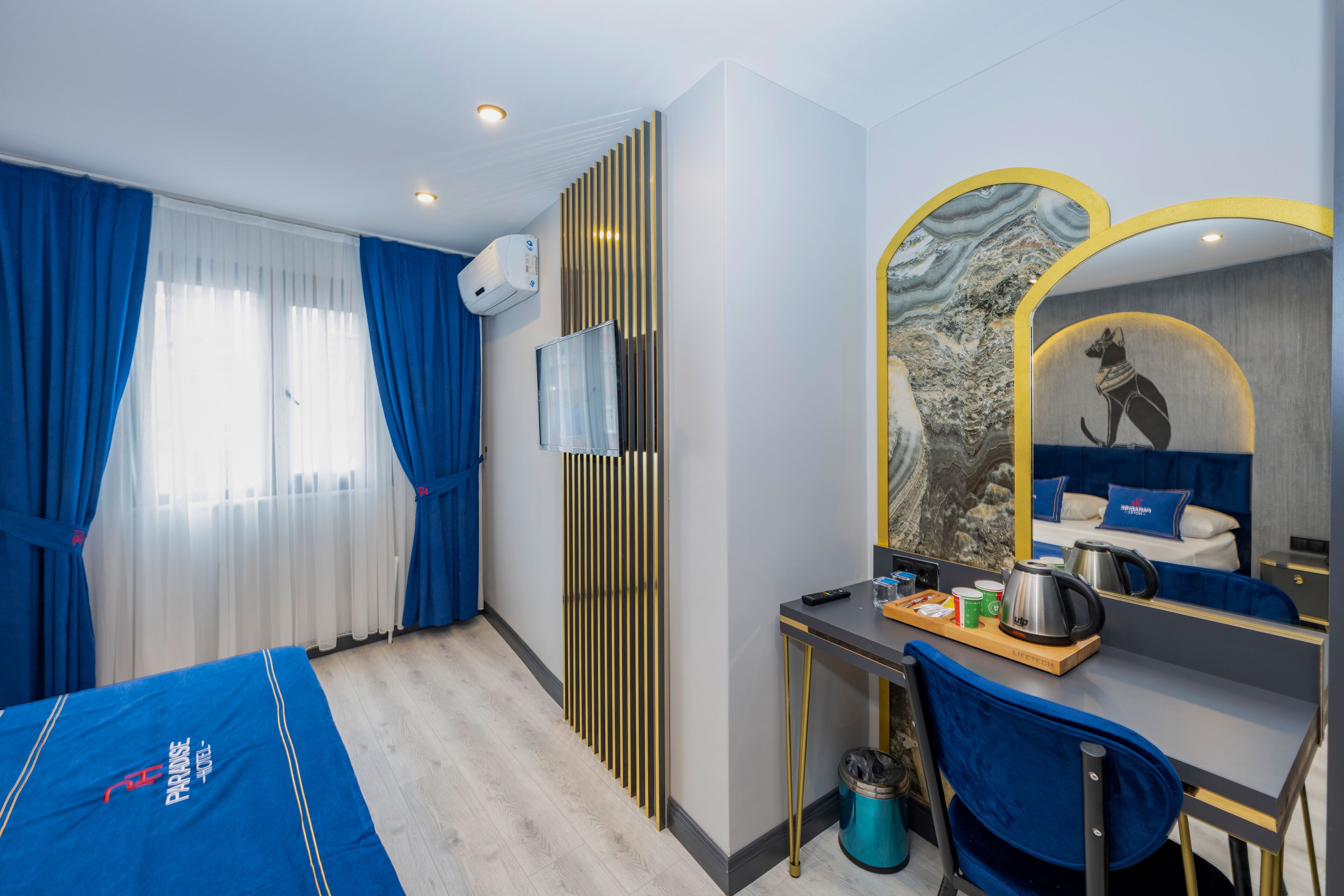 A hotel room with a bed, a desk with a chair, a mirror, and a window with curtains.