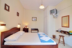 Una cama bien hecha con una toalla azul, una mesita con una lámpara, un televisor montado en la pared y una estantería con adornos.