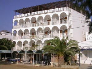 Un hotel de varios pisos con balcones, mobiliario exterior y una zona cubierta con barcos.