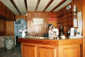 Un mostrador de recepción de madera con un hombre hablando por teléfono, un jarrón con flores y un tablón de anuncios.