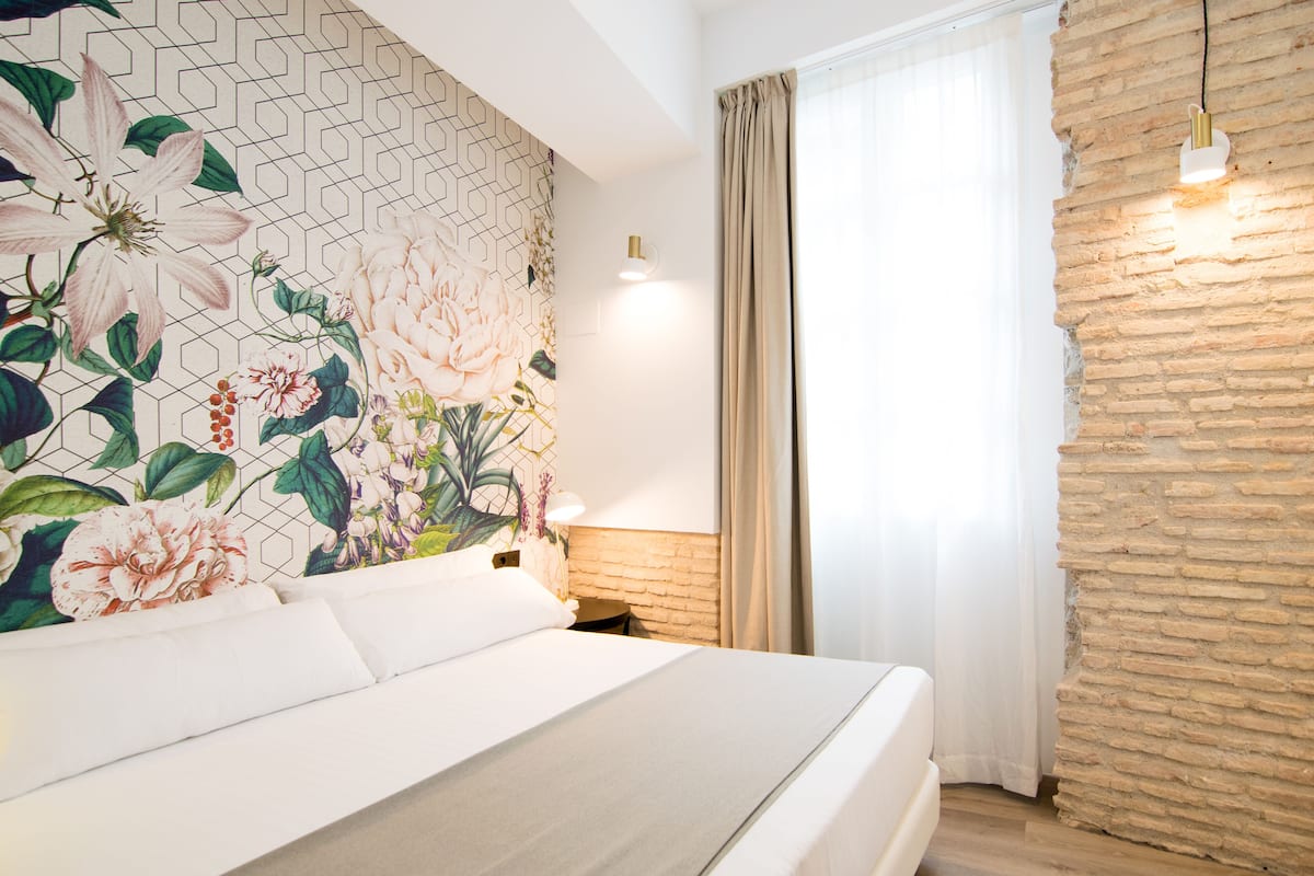 A neatly made bed with white linens, a floral-patterned wall, a brick accent wall, and a hanging light fixture.