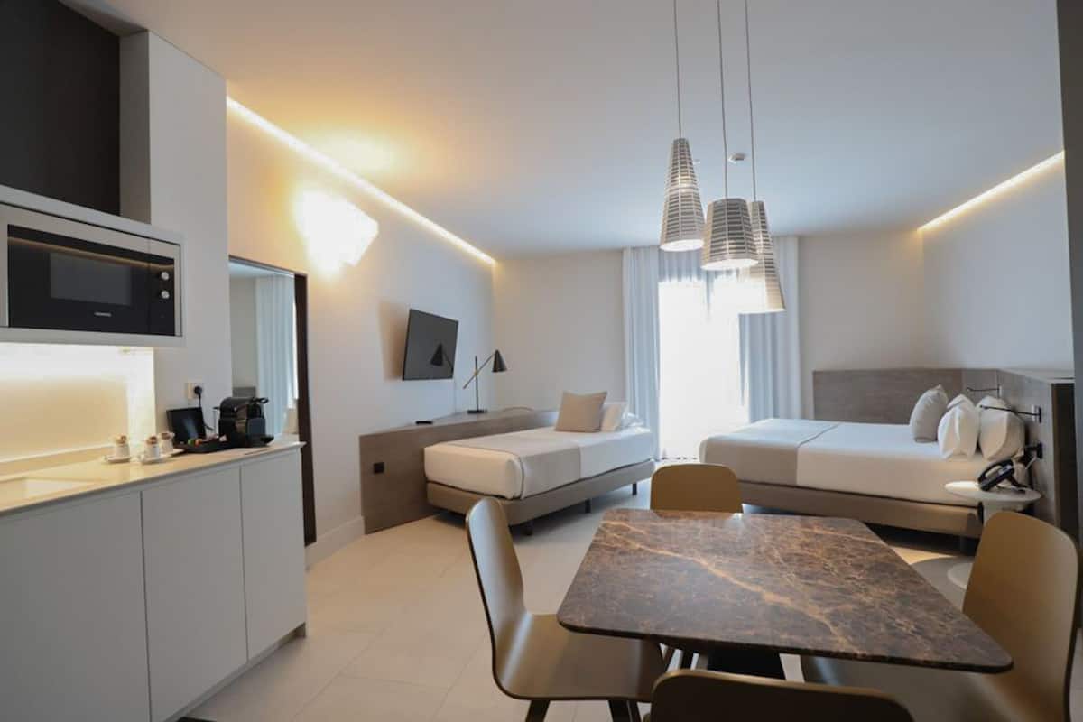 A modern, compact hotel room with a kitchenette, a dining area, a sofa bed, and a TV.