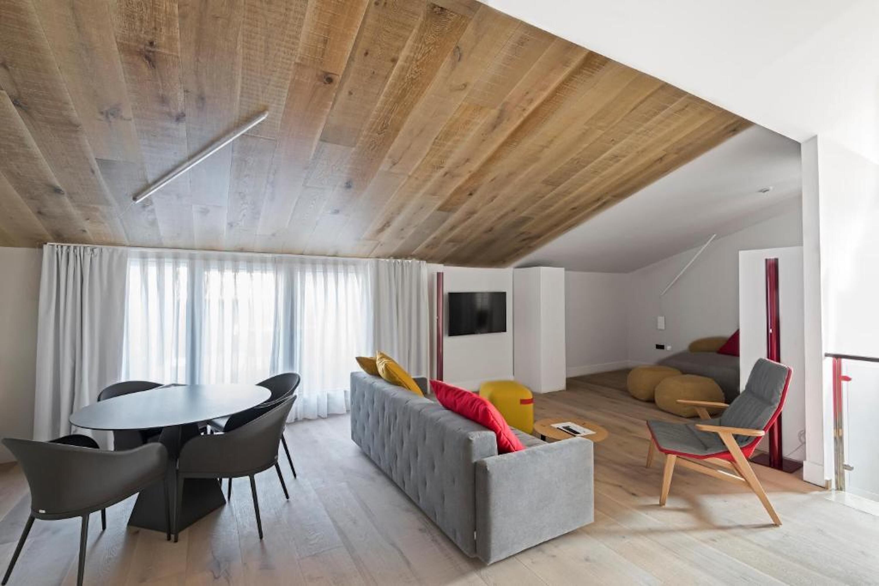Un salon moderne comprenant un coin repas, un canapé gris, un plafond en bois et un téléviseur à écran plat fixé au mur.