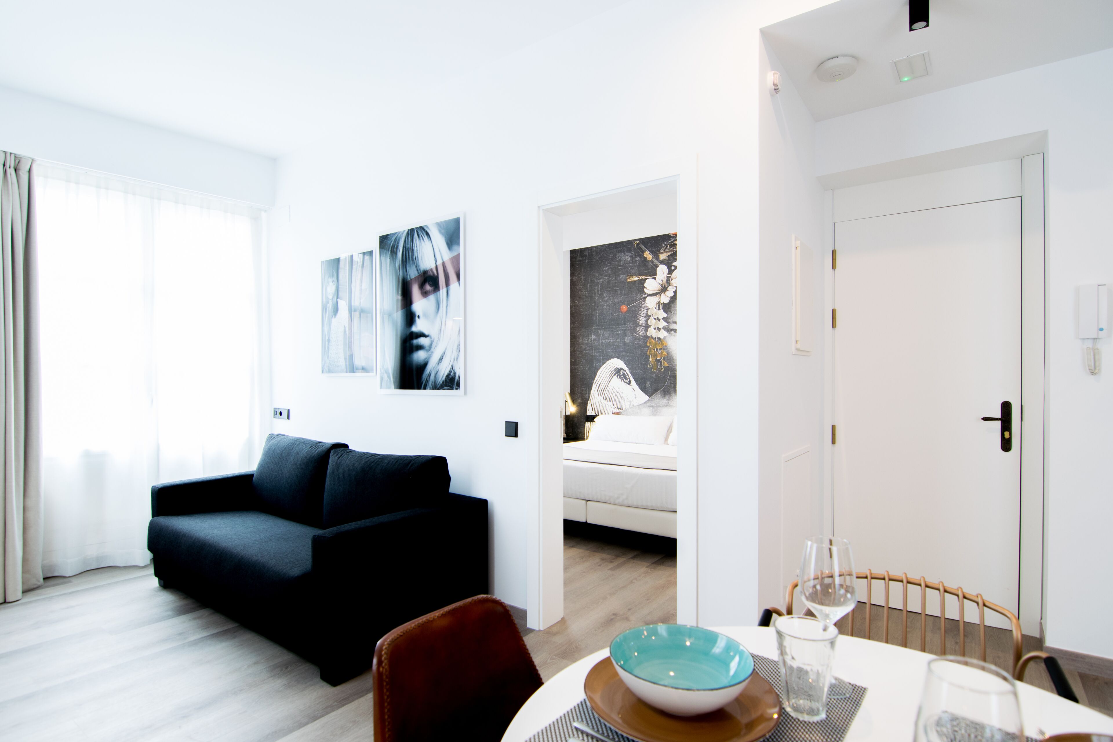 A modern living room with a dark sofa, a dining table with a bowl and wine glasses, and a bedroom visible through an open door.