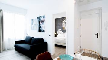 A modern living room with a dark sofa, a dining table with a bowl and wine glasses, and a bedroom visible through an open door.