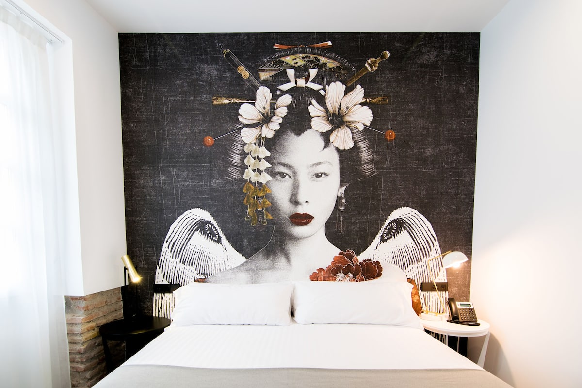 A bedroom with a large tapestry on the wall, a bed with white linens, a bedside table with a lamp and a phone, and a brick accent wall.