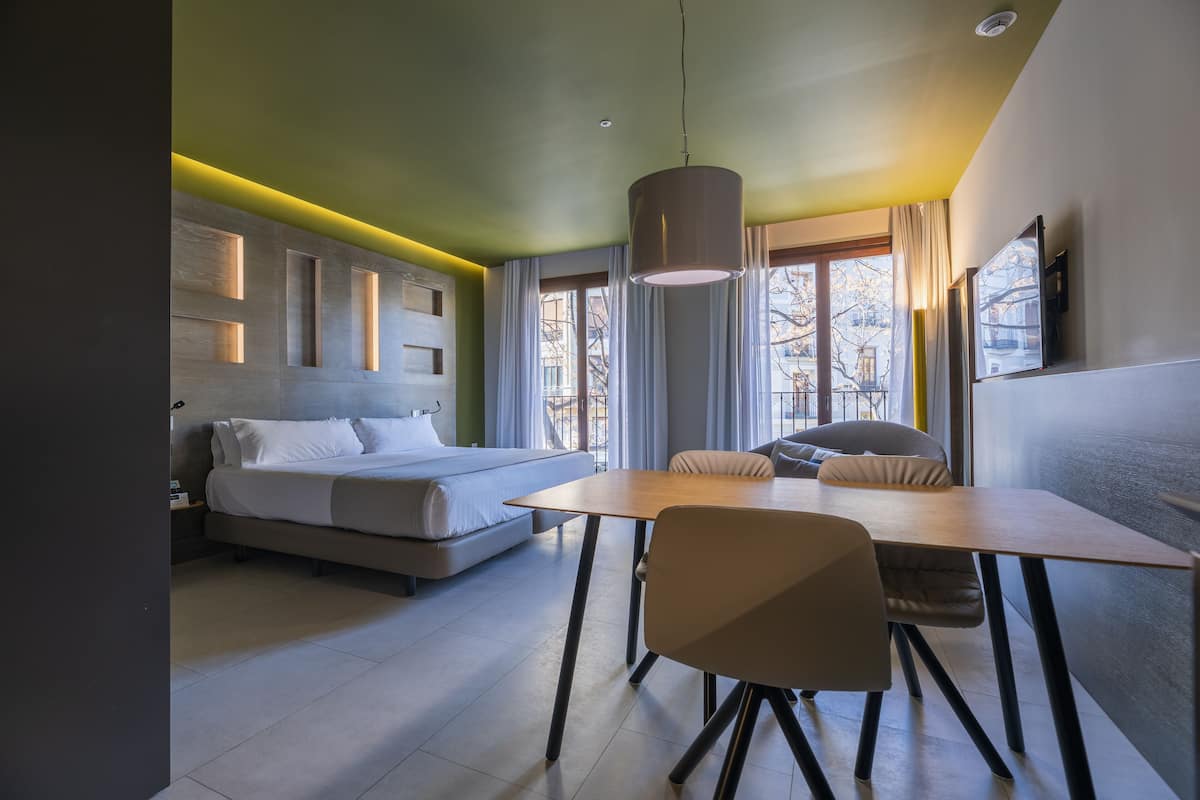 A modern hotel room with a bed, a dining table, and a chair. There is a window with curtains and a view of the outside.