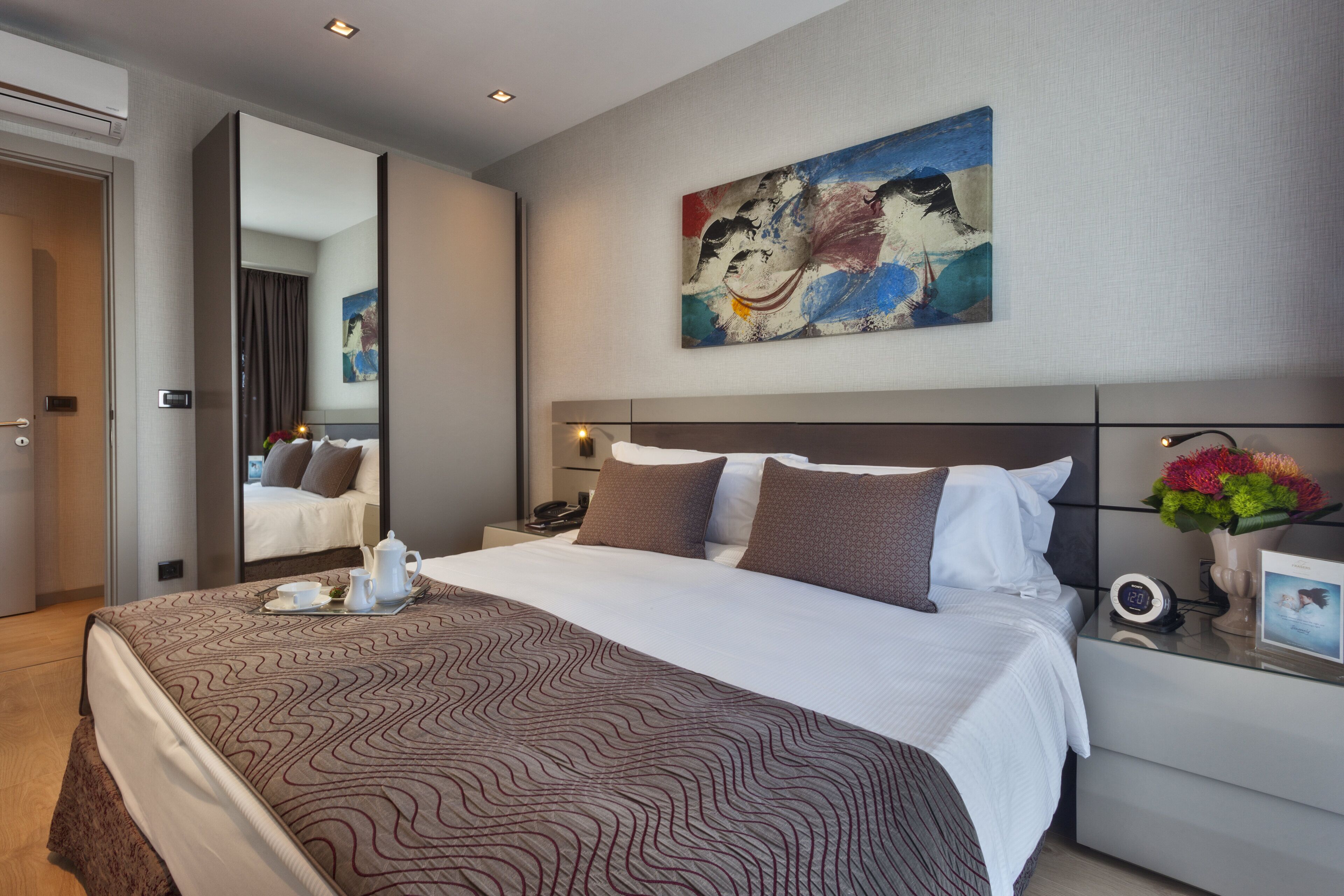 A modern hotel room with a large bed, a bedside table with a lamp, a painting on the wall, and a view of the adjacent room.