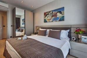 A modern hotel room with a large bed, a bedside table with a lamp, a painting on the wall, and a view of the adjacent room. - Antasya Suites (Istanbul)