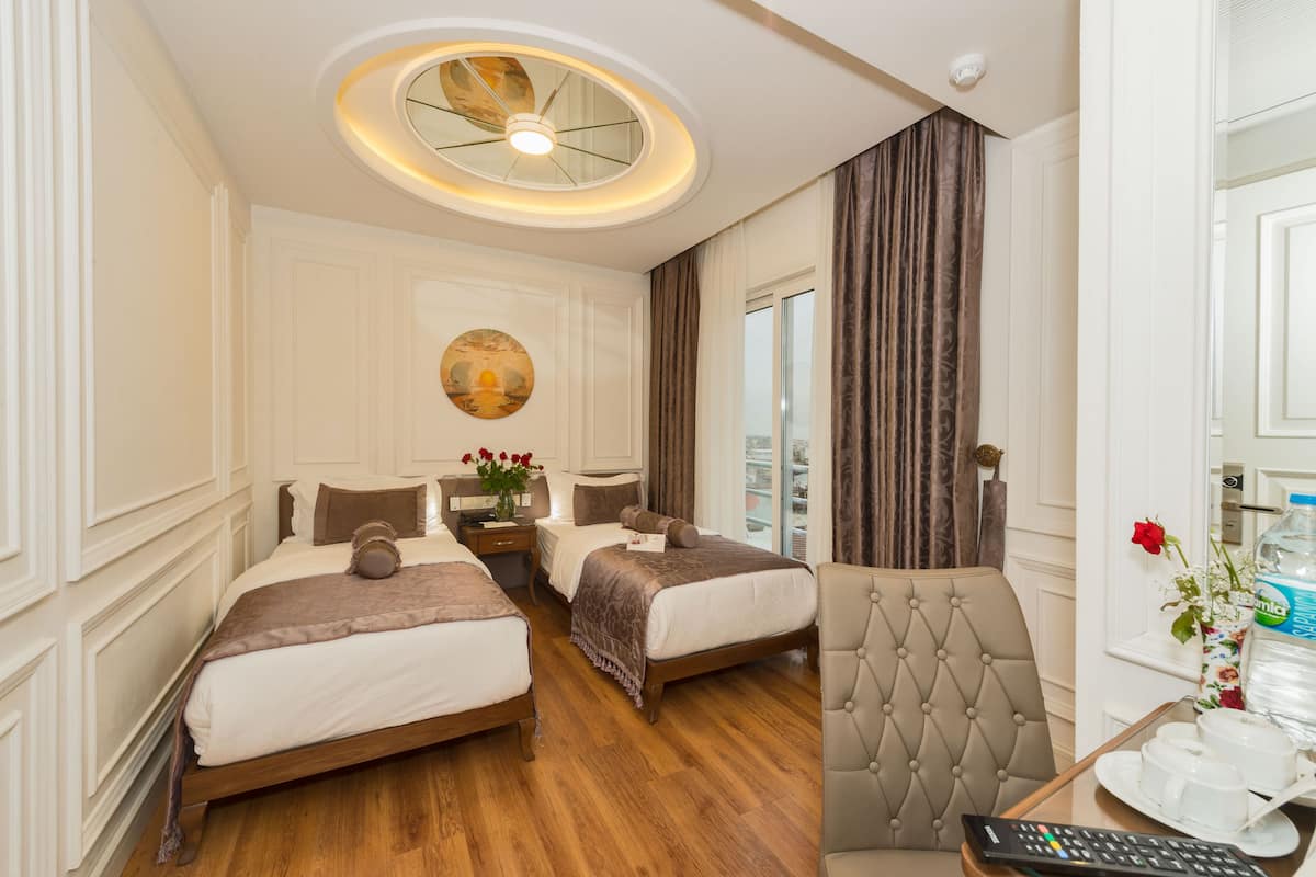 A hotel room with two beds, a round ceiling light, a painting on the wall, and a view of the city through the window.