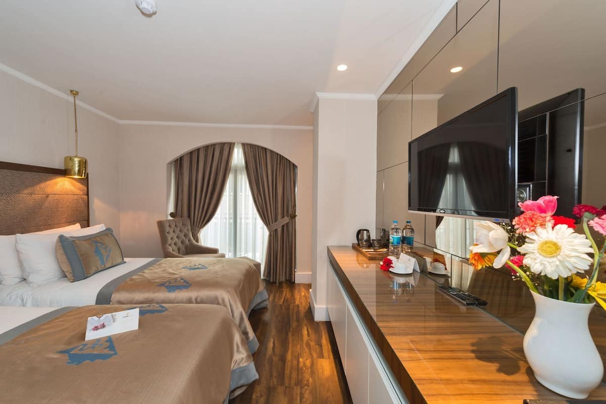 A hotel room with a bed, a chair, a desk with a TV, and a vase with flowers.