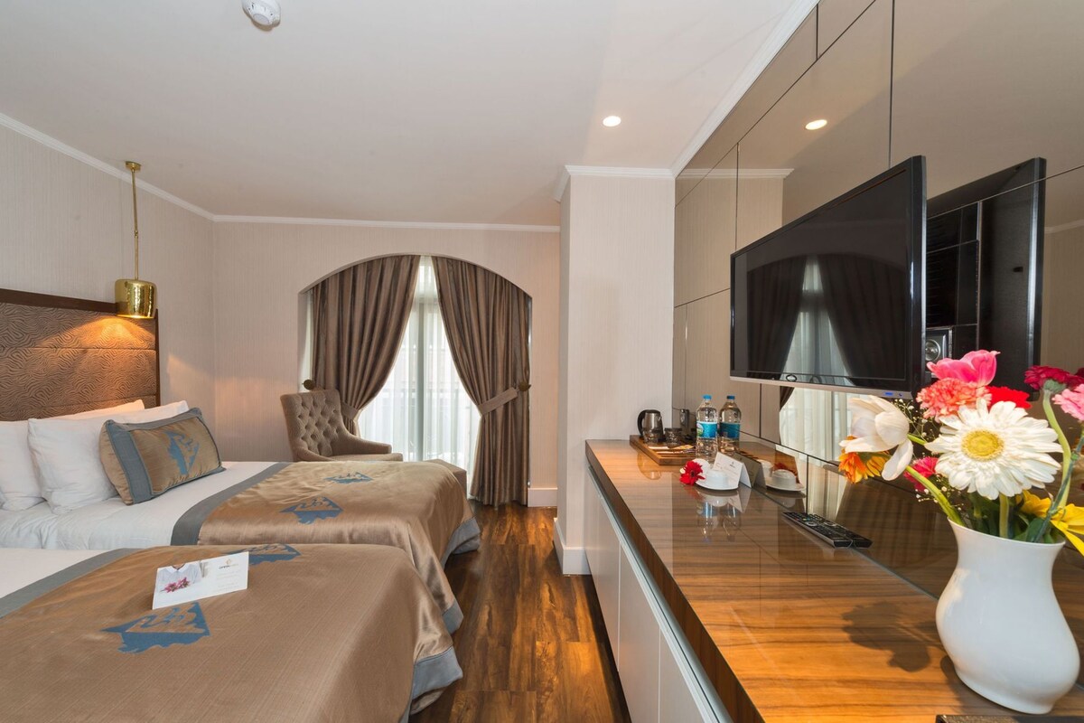 A hotel room with a bed, a chair, a desk with a TV, and a vase with flowers.