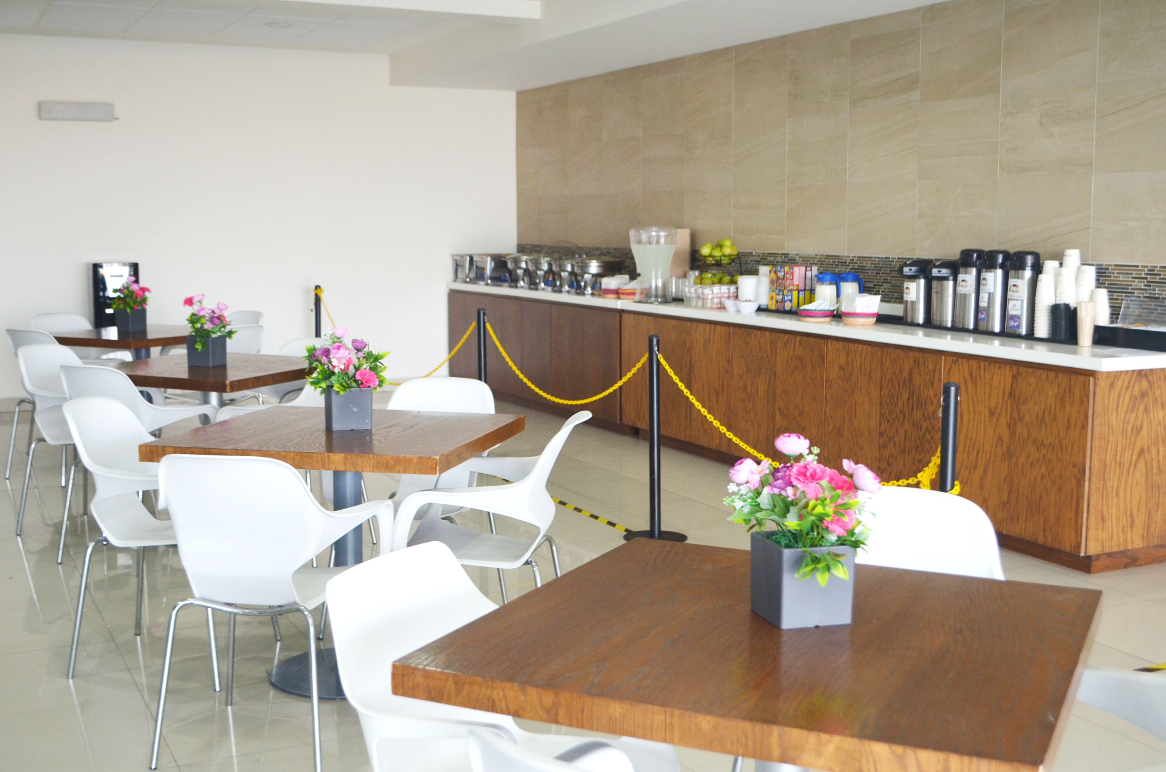 A modern dining area with wooden tables and white chairs, a kitchen counter with various appliances, and yellow ropes indicating a restricted area.