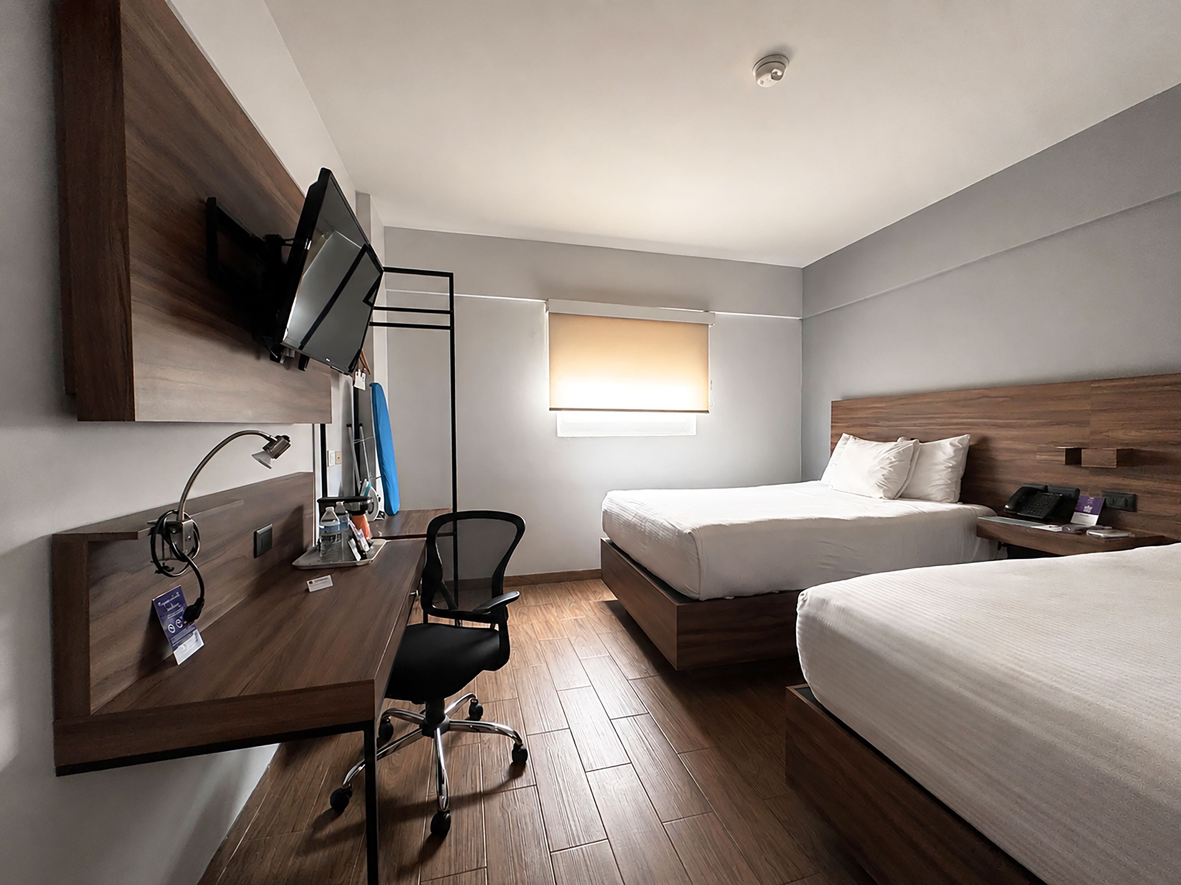 Habitación de hotel con escritorio, silla, dos camas, televisor montado en la pared y ventana con persianas.