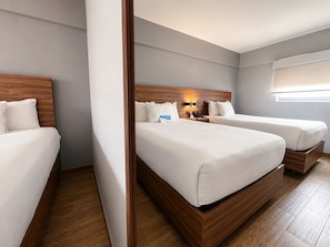 Quarto de hotel com duas camas, uma cabeceira de madeira, mesa de cabeceira com abajur e janela com persianas.