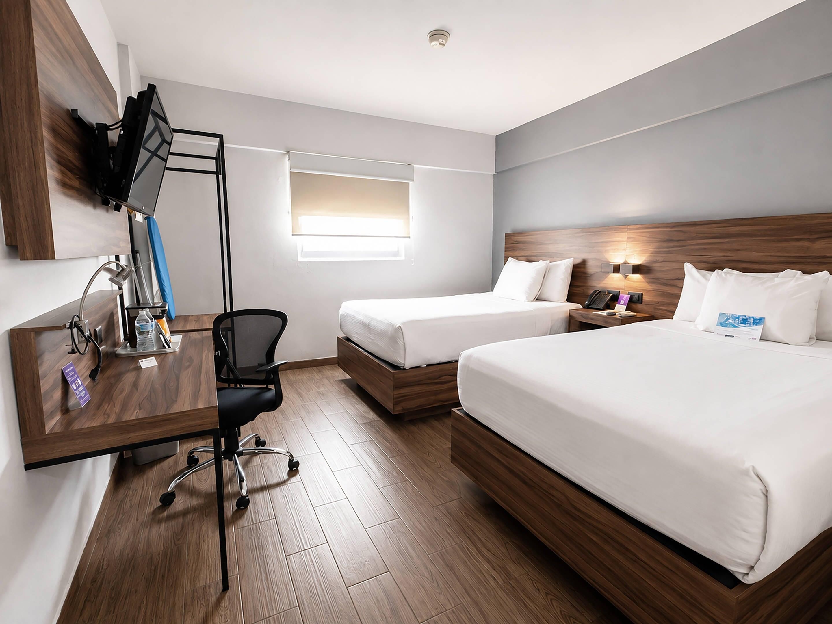 A hotel room with two beds, a desk, a chair, a TV, and a window.
