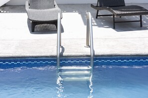 A swimming pool with metal handrails, two wicker lounge chairs, and a tiled edge.