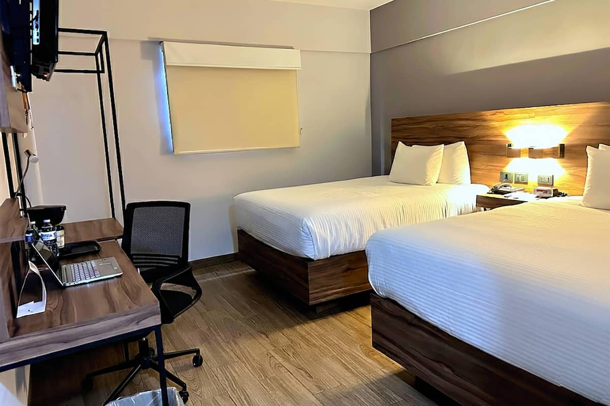 A hotel room with two beds, a desk with a laptop, a chair, a TV, and a bedside table with a lamp.