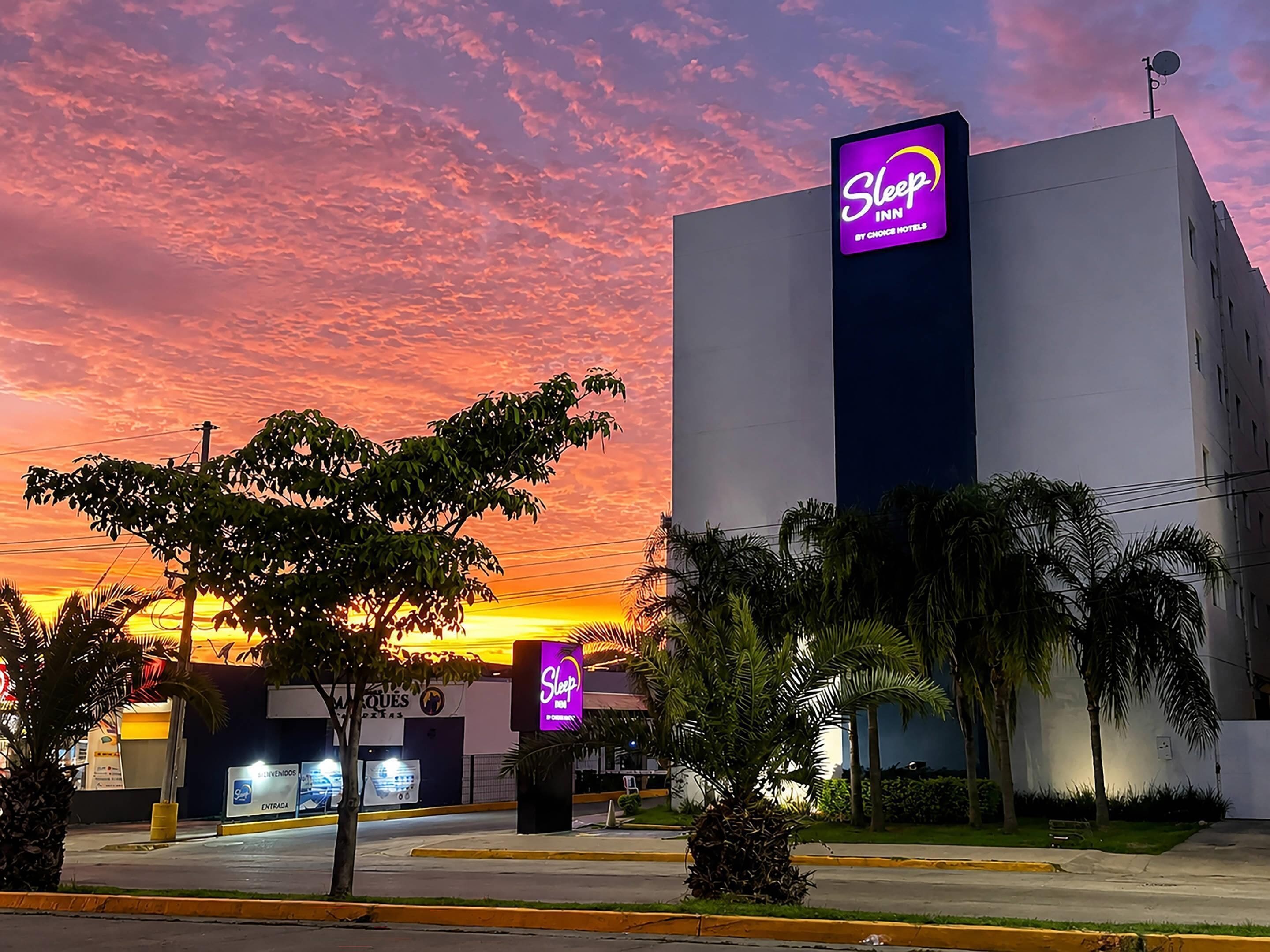 Um hotel Sleep Inn com uma placa grande, cercado por palmeiras e um céu ao pôr do sol.