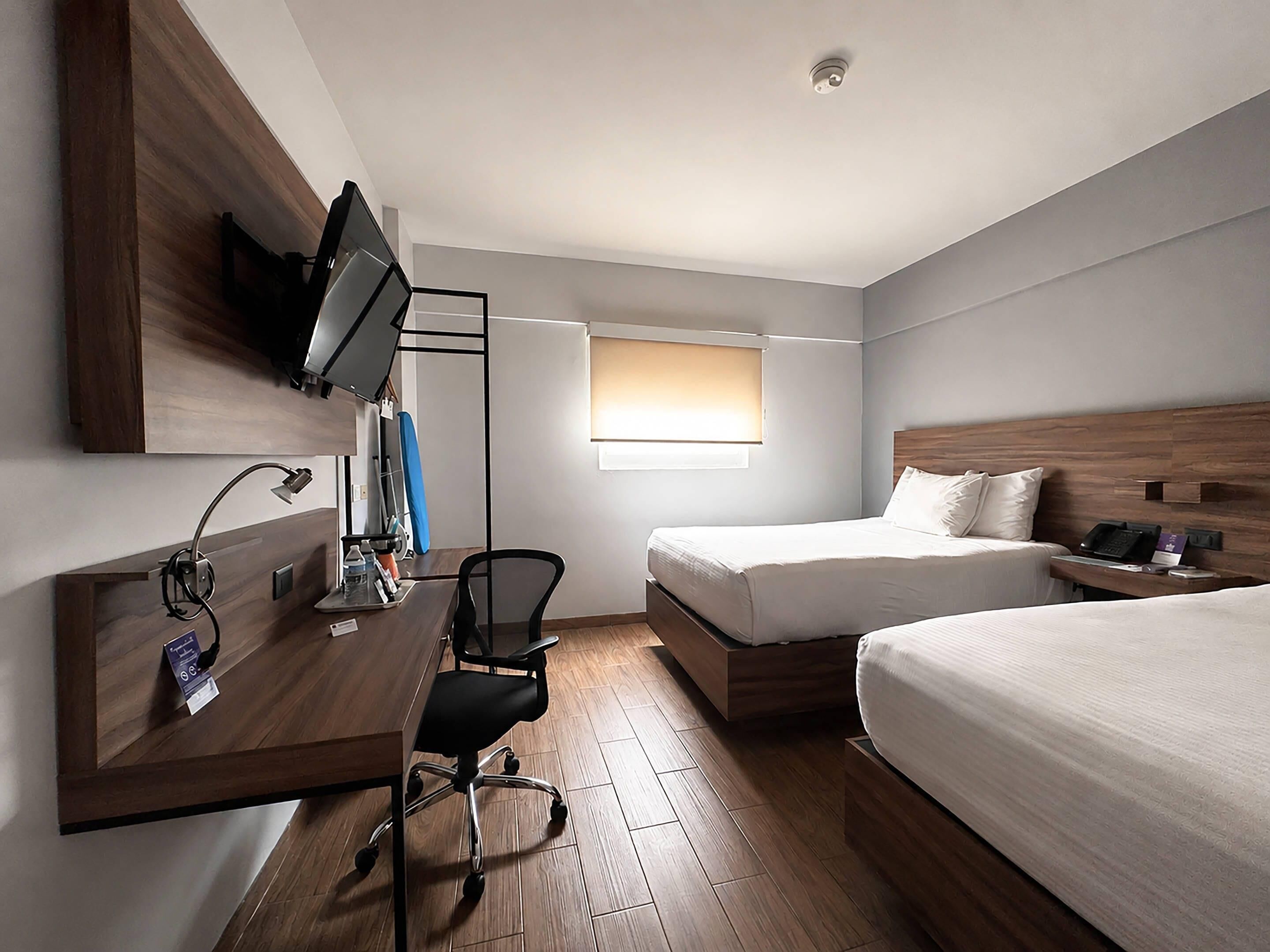 A hotel room with a desk, chair, two beds, a TV mounted on the wall, and a window with blinds.