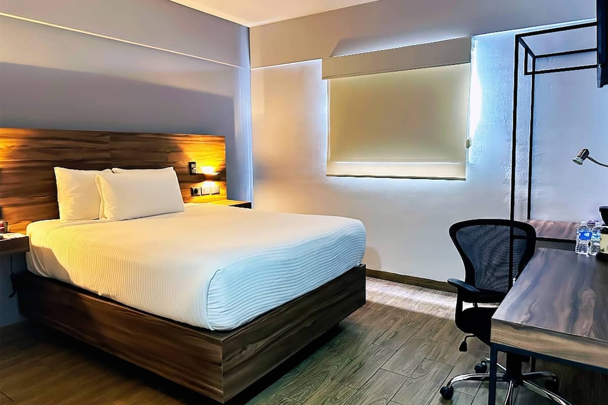 A hotel room with a wooden bed, a desk with a chair, a window, and a wall-mounted lamp.