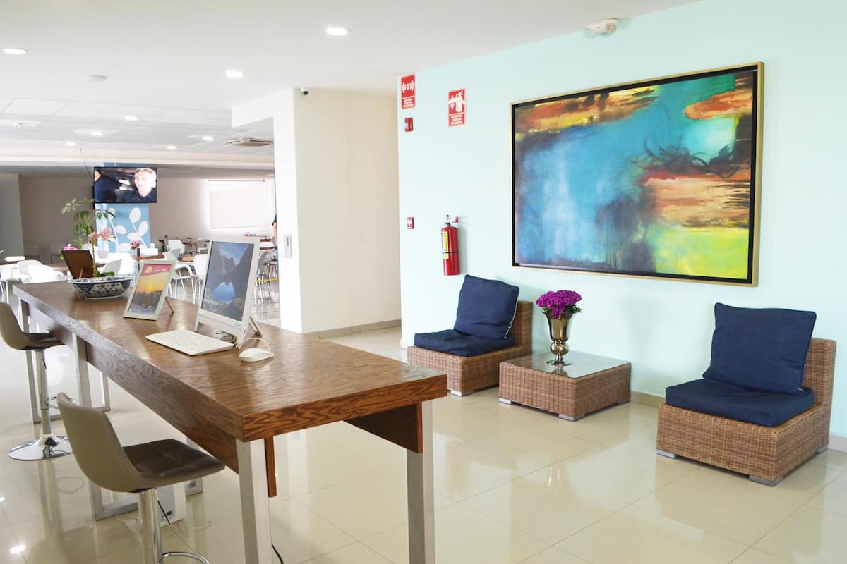 A reception area with a long wooden reception desk, two wicker armchairs with blue cushions, a large abstract painting on the wall, and a fire extinguisher mounted on the wall.