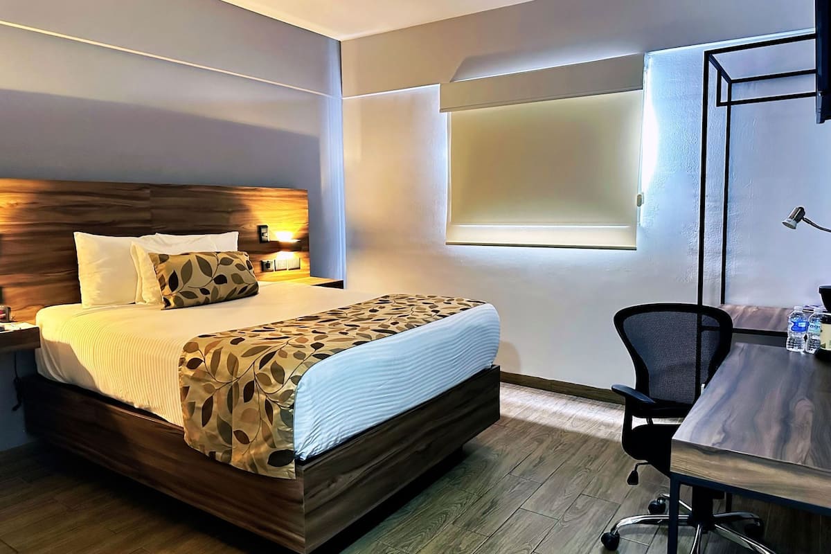 A hotel room with a bed, bedside table, lamp, desk, chair, and a window.