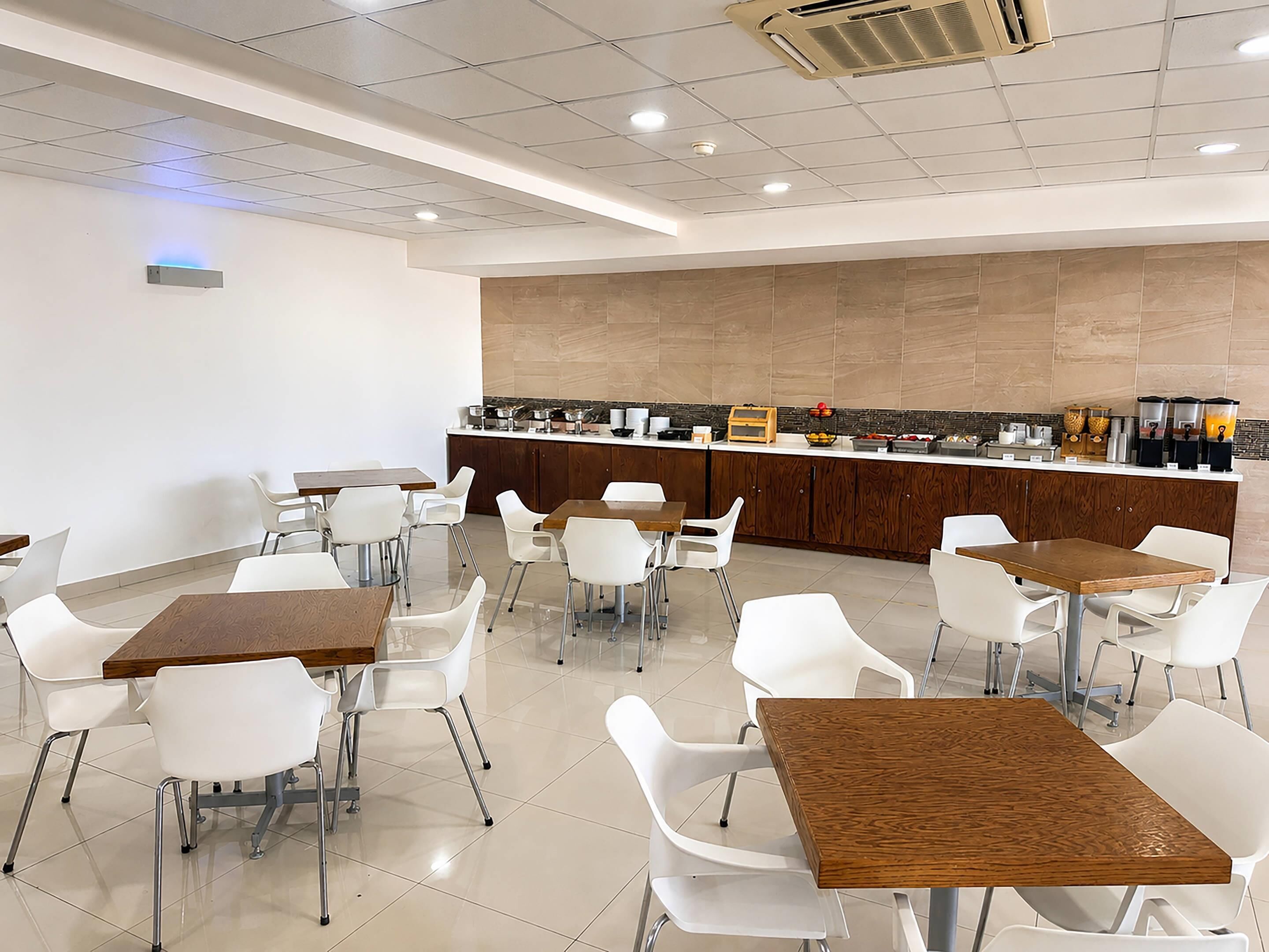 A modern dining area with white chairs and wooden tables, a kitchen counter with various appliances, and a ceiling with recessed lighting.