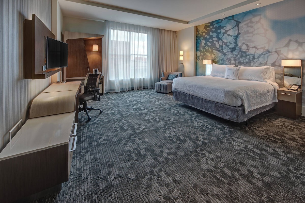 A hotel room with a large bed, a desk, a chair, a TV, and a painting on the wall.