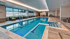 A large indoor swimming pool with clear lane markings, a seating area with cushioned chairs, and a view of the outside through large windows.